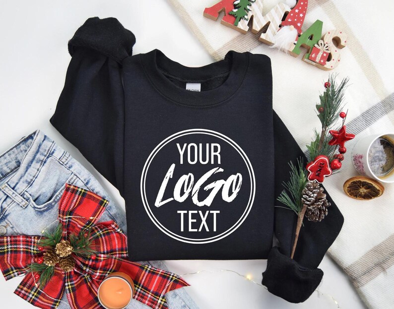 May include: A black crewneck sweatshirt featuring a white circular graphic on the chest with the text "YOUR LOGO TEXT" printed in a bold, sans-serif font. The sweatshirt has long sleeves and a ribbed collar, cuffs, and hem.