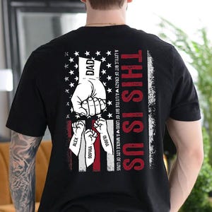 May include: Black t-shirt with a graphic design featuring an American flag motif and the words "THIS IS US." The design includes a hand holding three smaller fists, with the names ALEX, SOPHIA, and OLIVIA. The word "DAD" is also present.