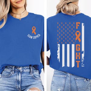 May include: Royal blue t-shirt with an orange awareness ribbon and the text "Team Sophia" on the front. The back features an orange ribbon design resembling a flag, with the word "FIGHT" in orange.