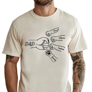 May include: Cream-colored t-shirt featuring a black graphic of a fist bump with the word "DAD" and smaller fist bumps labeled "David", "Olivia", and "Sophia". A paw print with the name "Rocky" is also included.