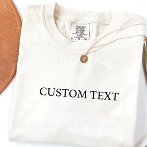 May include: A cream-colored short-sleeve t-shirt featuring the black text CUSTOM TEXT printed on the front. The shirt has a crew neckline and a Comfort Colors brand label at the inner collar, accompanied by a thin gold-toned chain necklace.