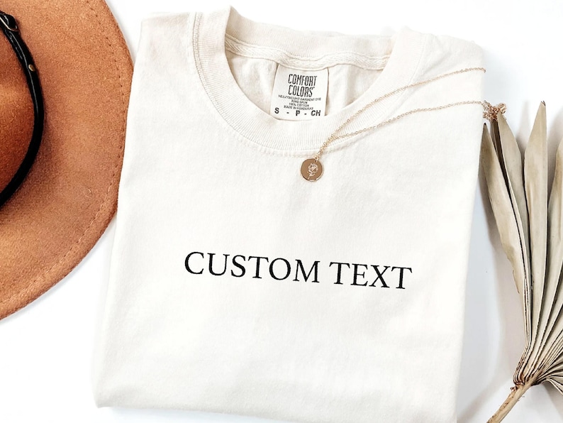 May include: A cream-colored short-sleeve t-shirt featuring the black text CUSTOM TEXT printed on the front. The shirt has a crew neckline and a Comfort Colors brand label at the inner collar, accompanied by a thin gold-toned chain necklace.