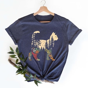 May include: Navy blue t-shirt featuring a cat silhouette filled with a forest scene. The cat's body is filled with trees, flowers, and a sunset. The shirt is made of a soft material and has rolled-up sleeves.