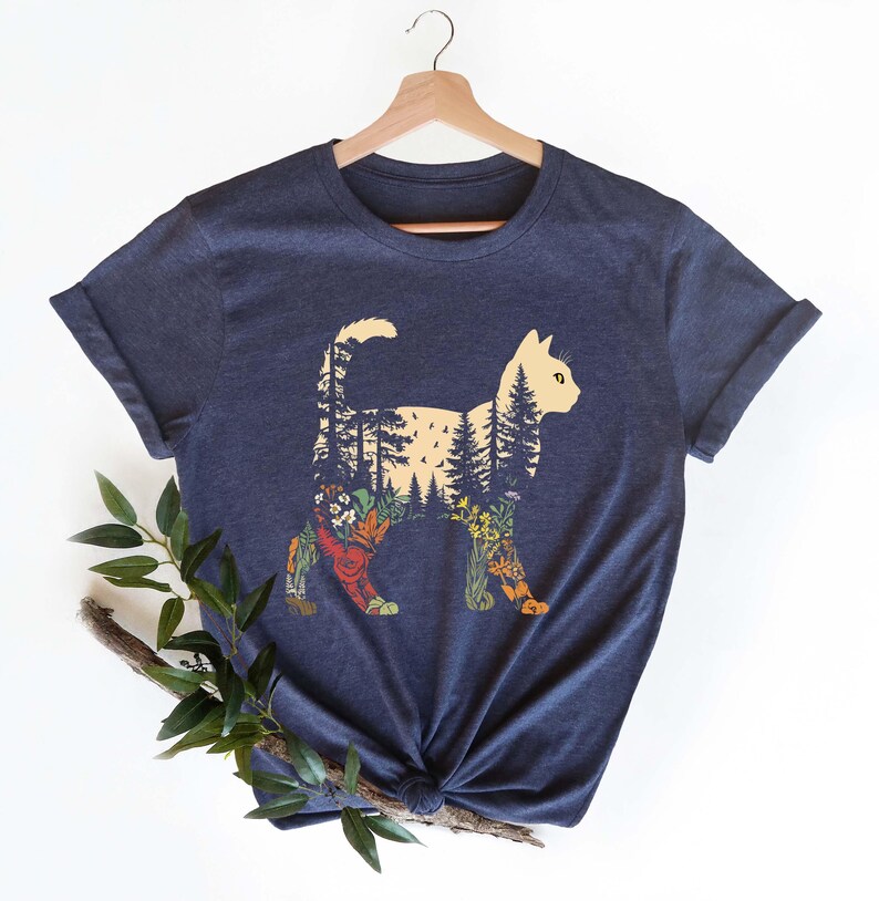 May include: Navy blue t-shirt featuring a cat silhouette filled with a forest scene. The cat's body is filled with trees, flowers, and a sunset. The shirt is made of a soft material and has rolled-up sleeves.