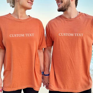 May include: Two matching short-sleeved t-shirts in a solid burnt orange color. Each shirt features the words CUSTOM TEXT printed in a simple white sans-serif font across the center of the chest. The shirts have a crew neckline and a relaxed fit.