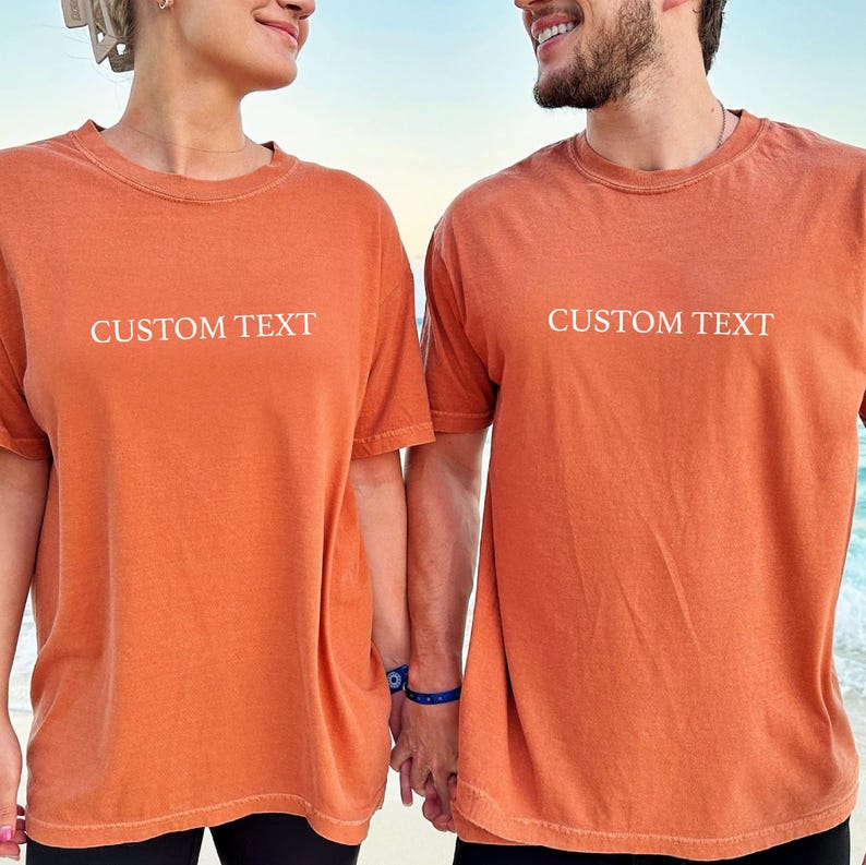 May include: Two matching short-sleeved t-shirts in a solid burnt orange color. Each shirt features the words CUSTOM TEXT printed in a simple white sans-serif font across the center of the chest. The shirts have a crew neckline and a relaxed fit.