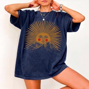 May include: Navy blue oversized t-shirt featuring a sun graphic with a smiling face and mountain range. The sun design is in shades of gold and orange. The shirt has short sleeves and a relaxed fit.