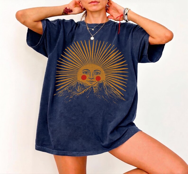 May include: Navy blue oversized t-shirt featuring a sun graphic with a smiling face and mountain range. The sun design is in shades of gold and orange. The shirt has short sleeves and a relaxed fit.