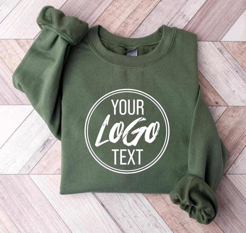 May include: An olive green crewneck sweatshirt featuring a white circular graphic on the chest. Inside the double-lined circle, the text reads YOUR LOGO TEXT in a combination of clean sans-serif and stylized brush-stroke lettering.