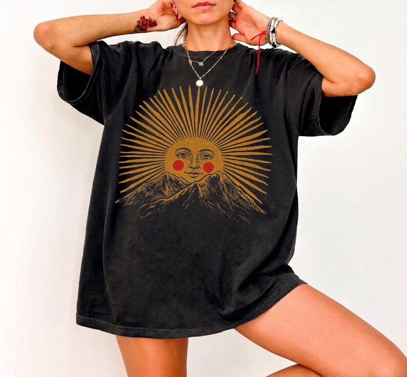 May include: Black t-shirt featuring a sun graphic with a face, radiating lines, and mountain imagery. The sun is in shades of gold and the face has red cheeks. The shirt has short sleeves and a relaxed fit.
