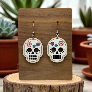 May include: A pair of wooden skull earrings with a floral design. The earrings feature a white skull shape with black eye sockets and a mouth, and are adorned with pink, purple, and green flowers. The earrings are displayed on a wooden stand.