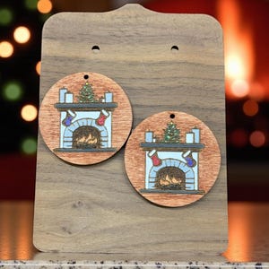 以下が含まれることがあります： クリスマスをテーマにした木製の丸いイヤリング。各イヤリングは、暖炉、靴下、クリスマスツリーの詳細なイラストが特徴です。イヤリングは木製のスタンドに展示されており、お祝いの機会に最適です。