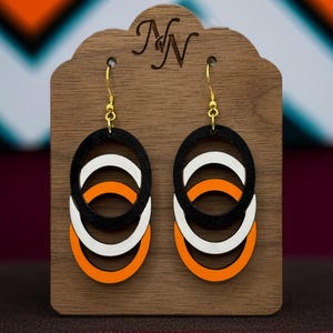 Puede incluir: Pendientes de madera con diseño ovalado en capas en negro, blanco y naranja. Los pendientes cuelgan de ganchos dorados y se exhiben en un soporte de madera con las iniciales "NN" grabadas.