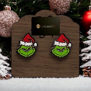 Puede incluir: Pendientes de botón con temática del Grinch. Cada pendiente presenta la cara del Grinch con un gorro de Papá Noel rojo y ribete blanco. Los pendientes se exhiben en una tarjeta de madera con el logotipo de LuxLaserCo, rodeados de adornos navideños.