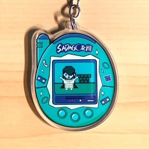 May include: A teal and white Sadagotchi keychain with a silver keyring. The keychain features a pixelated character on the screen. The text "Sadagotchi" is displayed at the top of the screen. The keychain is made of clear acrylic.