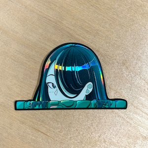 May include: A holographic sticker featuring a cartoon illustration of a woman with long dark blue hair peeking over a green and blue patterned border.