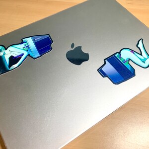 K&ouml;nnte beinhalten: Zwei Aufkleber auf einem silbernen Laptop. Die Aufkleber zeigen eine Frau in einem blauen und gr&uuml;nen Outfit mit einem Computermonitor. Die Frau liegt in einer liegenden Position.