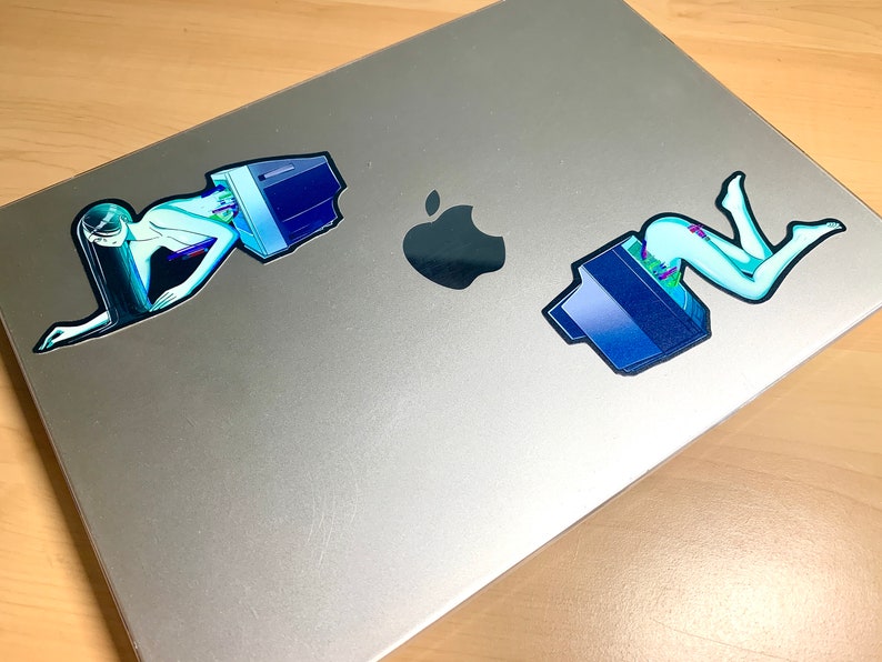 K&ouml;nnte beinhalten: Zwei Aufkleber auf einem silbernen Laptop. Die Aufkleber zeigen eine Frau in einem blauen und gr&uuml;nen Outfit mit einem Computermonitor. Die Frau liegt in einer liegenden Position.