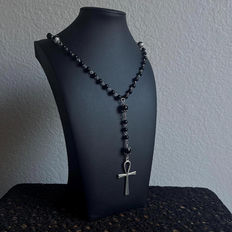May include: A black beaded necklace with a silver ankh pendant. The necklace is displayed on a black mannequin bust.