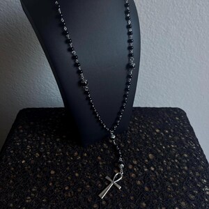 May include: A black beaded necklace with a silver ankh pendant. The necklace is on a black display stand.