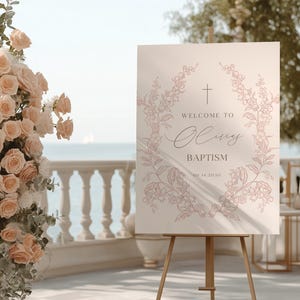 Pode incluir: Um letreiro de boas-vindas para uma cerimónia de batismo. O letreiro, sobre um cavalete de madeira, apresenta o texto "Welcome to Olivia Baptism" em uma escrita delicada, rodeado por uma coroa floral. Rosas cor-de-rosa e vegetação adornam o fundo.