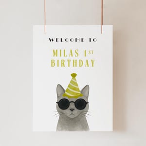 以下が含まれることがあります： 「WELCOME TO MILAS 1ST BIRTHDAY」の文字が黒と黄緑色で書かれた白いポスター。サングラスとパーティーハットをつけた灰色の猫の水彩イラストが下にあります。
