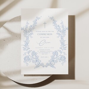 May include: A cream-colored invitation with blue floral wreath and cross design. The text reads "PLEASE JOIN US FOR THE COMMUNION OF OUR SON" with event details. The invitation is on a light gray surface.