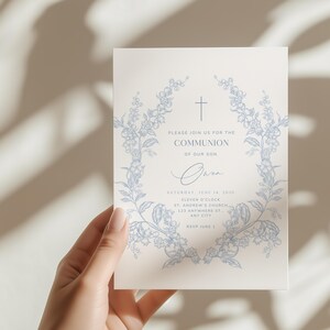 May include: A cream-colored invitation card with a blue floral wreath design and a cross at the top. The text reads "Please join us for the Communion of our son" with event details. The card is held by a hand.