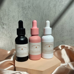 Puede incluir: Tres botellas de vidrio con tapas negras, rosadas y blancas. Cada botella tiene una etiqueta que dice "Scent from Heaven" con un pequeño icono de gota. Las botellas están dispuestas en una plataforma hexagonal de color marrón claro.