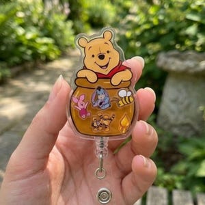 May include: A clear acrylic badge reel featuring a yellow Winnie the Pooh character resting on a honey pot. The honey pot is decorated with small illustrations of Piglet, Eeyore, Tigger, a bee, and a honey drop. The reel has a retractable cord.
