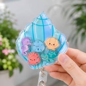 May include: A clear acrylic badge reel shaped like a large blue seashell, containing smaller floating charms of a purple seahorse, a blue sand dollar, a peach scallop shell, a red anemone, and a green sea turtle, attached to a clear plastic strap.