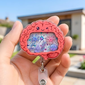 Könnte beinhalten: Ein rosa gehirnförmiger Ausweishalter mit einem zwinkernden Gesicht und einem zentralen Fenster, das mit bunten Illustrationen von Neuronen, DNA-Strängen und wissenschaftlichen Symbolen gefüllt ist. Das Acrylstück ist an einem transparenten Kunststoffband befestigt.