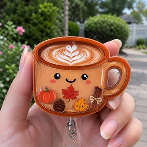 May include: An acrylic badge reel featuring a light orange coffee mug with a smiling face, latte art, and autumn-themed charms including a pumpkin, maple leaf, pinecone, small leaf, and cinnamon sticks attached to a clear plastic retractable cord.