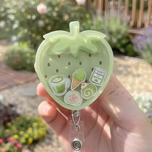 May include: A light green strawberry-shaped badge reel featuring a clear front filled with small floating charms including a matcha latte, an ice cream cone, a matcha jar, and a green roll cake. The badge reel has a clear plastic strap and metal clip.