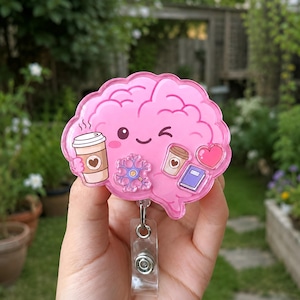 Könnte beinhalten: Ein gehirnförmiger Ausweishalter in Pink mit einem zwinkernden Gesicht, einer Kaffeetasse, einem Neuron, einer kleineren Kaffeetasse, einem Buch und einem rosa Herz. Das Acryl-Design hat eine glänzende Oberfläche und einen transparenten Clip.