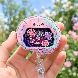 May include: A brain-shaped badge reel featuring a pastel-colored outer shell with a winking face. The center contains a dark purple, star-filled shaker compartment with small floating charms including a neuron, DNA strand, and virus icons.