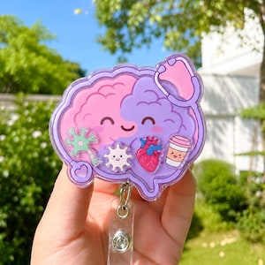 Könnte beinhalten: Ein Ausweishalter mit einem gehirnförmigen Acryl-Anhänger in einem rosa-violetten Farbverlauf. Das Gehirn hat ein lächelndes Gesicht und ist mit kleinen Symbolen eines Neurons, eines weißen Blutkörperchens, eines Herzens, eines Kaffees und eines Stethoskops verziert.