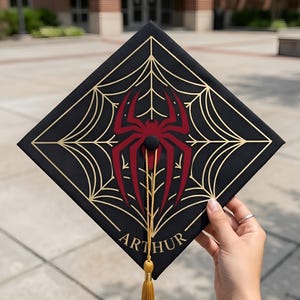 Könnte beinhalten: Schwarze Abschlusskappe mit einem goldenen Spinnennetz-Design und einem roten Spinnenemblem. Der Name "ARTHUR" ist in Gold aufgedruckt. Eine goldene Quaste hängt von der Mitte.