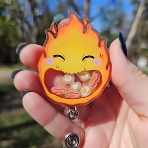May include: A badge reel featuring a smiling, cartoon-style flame character with a yellow and orange color scheme. The character's mouth is open, revealing small food-shaped charms. The reel has a silver clip and a retractable cord.