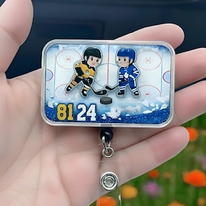 Könnte beinhalten: Rechteckige Ausweisrolle mit einem Hockey-Design, das zwei Cartoon-Hockeyspieler in gelben und blauen Uniformen zeigt. Die Zahl "8124" ist in gelber Schrift dargestellt. Die Rolle hat eine transparente Kunststoffabdeckung und eine einziehbare Schnur.