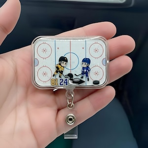 May include: A rectangular hockey-themed badge reel with a clear plastic cover. The design features two cartoon hockey players on an ice rink, a hockey puck, and the numbers "81 24". The reel has a silver-colored metal clip and a clear plastic strap.