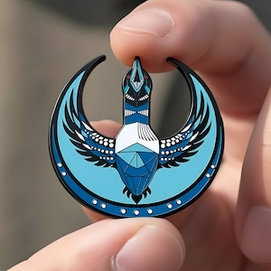 Pode incluir: Um alfinete redondo de esmalte com um desenho estilizado de pássaro em tons de azul, preto e branco. O pássaro está colocado contra uma forma de meia-lua. O alfinete é segurado entre dois dedos.