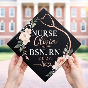 May include: A black graduation cap decorated with floral accents and a rose gold stethoscope design. The cap features the name "Olivia" and the title "NURSE" in white script, along with "BSN, RN 2026" and a heartbeat graphic.