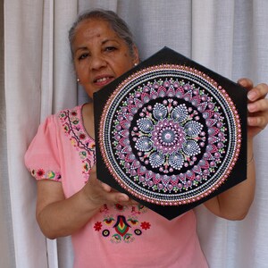 Op de afbeelding: Een zwart, zeshoekig canvas met een gedetailleerd mandala-ontwerp. De mandala bestaat uit roze, witte en zwarte stippen, gerangschikt in concentrische cirkels. Het kunstwerk wordt vastgehouden door een persoon die een roze geborduurd shirt draagt.