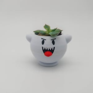 Op de afbeelding: Een witte keramische pot in de vorm van een spook met een rode tong en zwarte ogen. De pot is gevuld met een groene succulent.