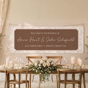 May include: A wedding banner with a brown rectangular center and a floral patterned border. The banner reads "WELCOME TO THE WEDDING OF Anne Hart & John Schofield MAY THIRTY FIRST • TWENTY TWENTY SIX". The banner is suspended above a table set for a wedding reception.