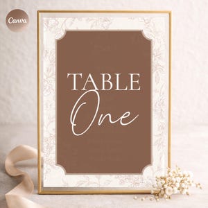 Puede incluir: Un letrero de mesa enmarcado con un centro marrón y un borde floral. El letrero dice "TABLE One" en una elegante escritura blanca. El texto adicional incluye "Bar" y una lista de cócteles. El marco es dorado.