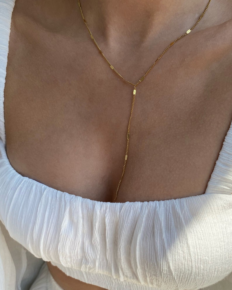 Op de afbeelding: Een delicate goudkleurige Y-vormige ketting. De ketting heeft een fijne ketting met kleine rechthoekige en ovale schakels. De hanger hangt naar beneden, wat een stijlvolle en elegante uitstraling cre&euml;ert. De ketting wordt getoond tegen een witte top.