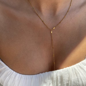 Op de afbeelding: Een delicate goudkleurige Y-vormige ketting. De ketting heeft een fijne ketting met kleine rechthoekige bedels. De ketting wordt gedragen over een witte top.