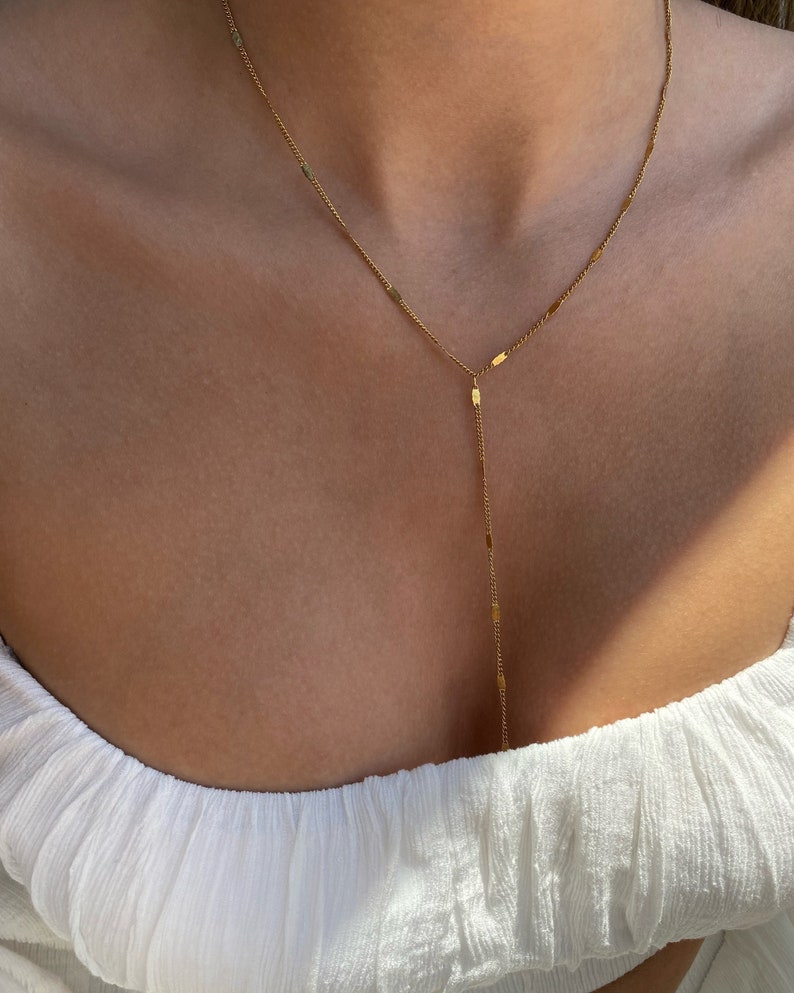 Op de afbeelding: Een delicate goudkleurige Y-vormige ketting. De ketting heeft een fijne ketting met kleine rechthoekige bedels. De ketting wordt gedragen over een witte top.